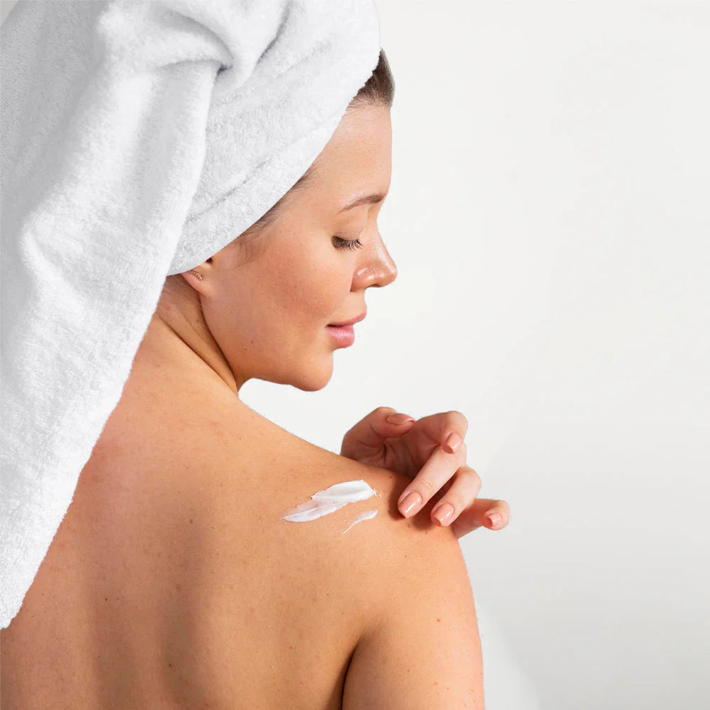 Woman applying cream to her shoulder with a white towel on her head against a light background